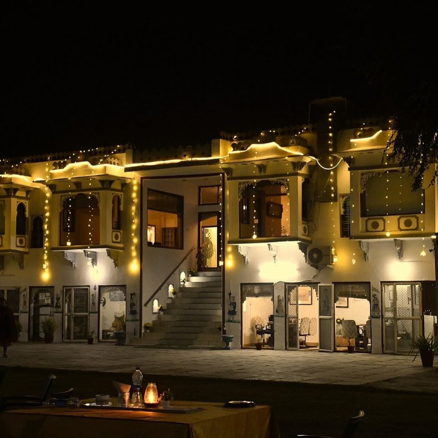 The front of the house at night, strung end to end with fairy lights, with a candle lantern on a laid table in the foreground.