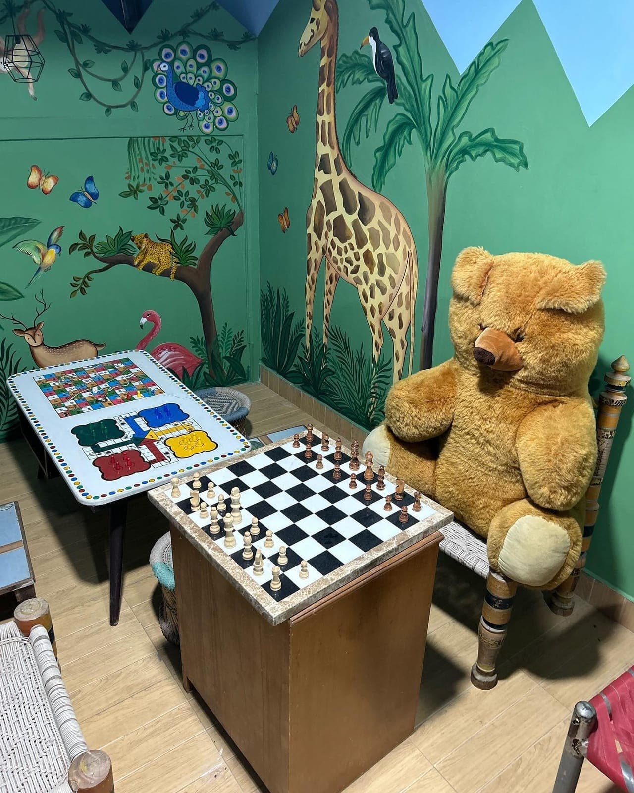 The games room, its walls painted with a giraffe and a peacock, a chess board and a ludo table set out and a large teddy bear in a chair.