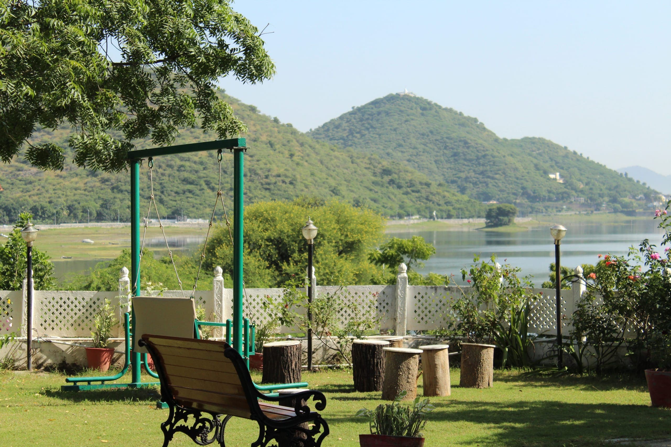 A green garden swing on the lawn, with Fateh Sagar and the Aravalli hills beyond the low white boundary wall.