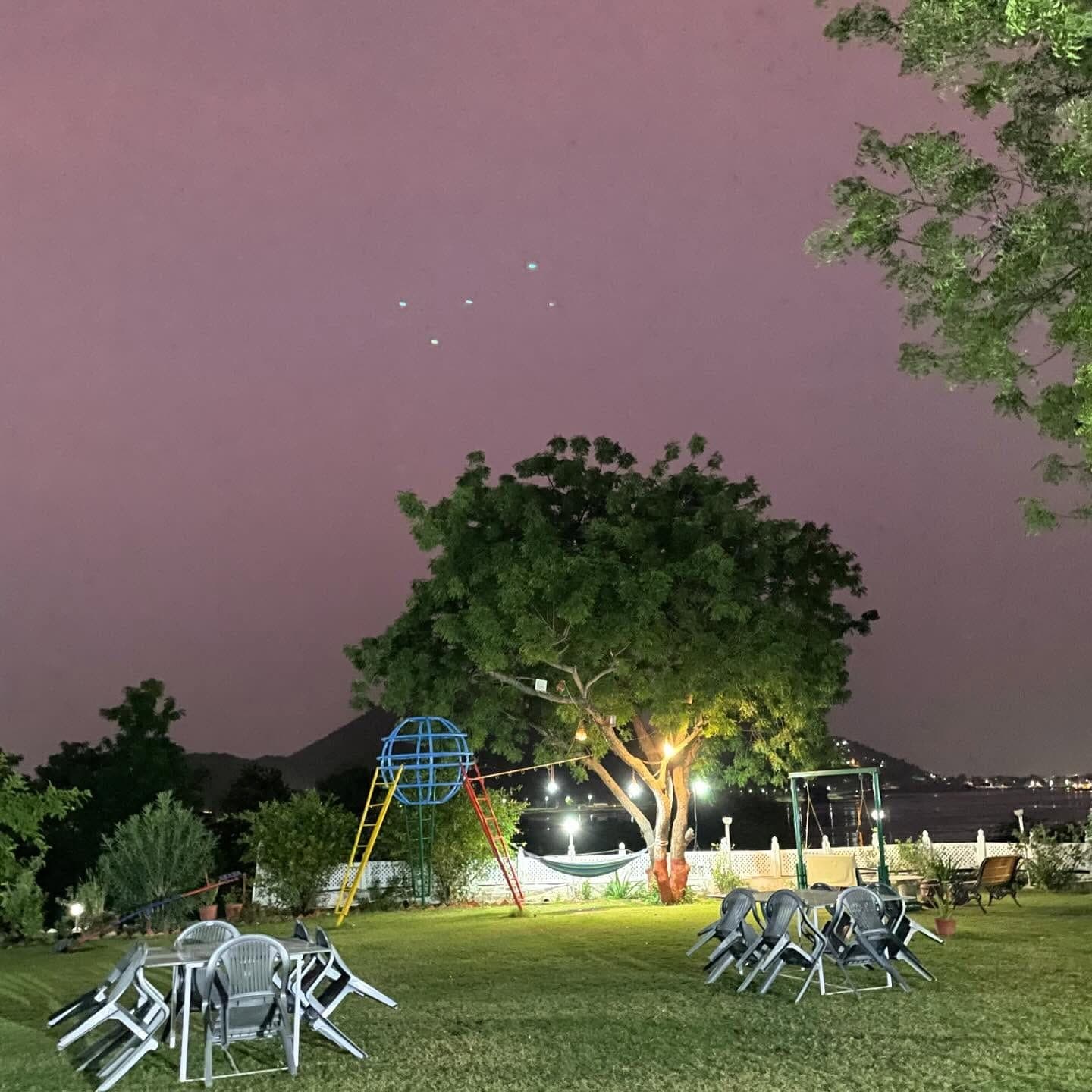 The lawn after dark with two tables and stacked garden chairs on the grass, a tree hung with bulbs, a hammock, and the lights of the far shore of Fateh Sagar beyond the wall