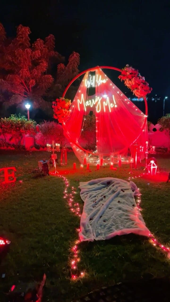 A lit circular arch draped in tulle on the night lawn, carrying a neon Will You Marry Me sign, with candles, flowers and a line of fairy lights leading across the grass to a rug