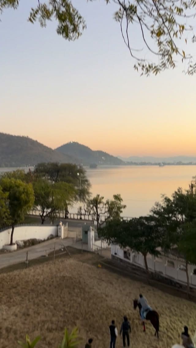 A rider on a dark horse crossing a dry slope at dawn, with Lake Fatehsagar and the Aravalli hills flat and pale behind