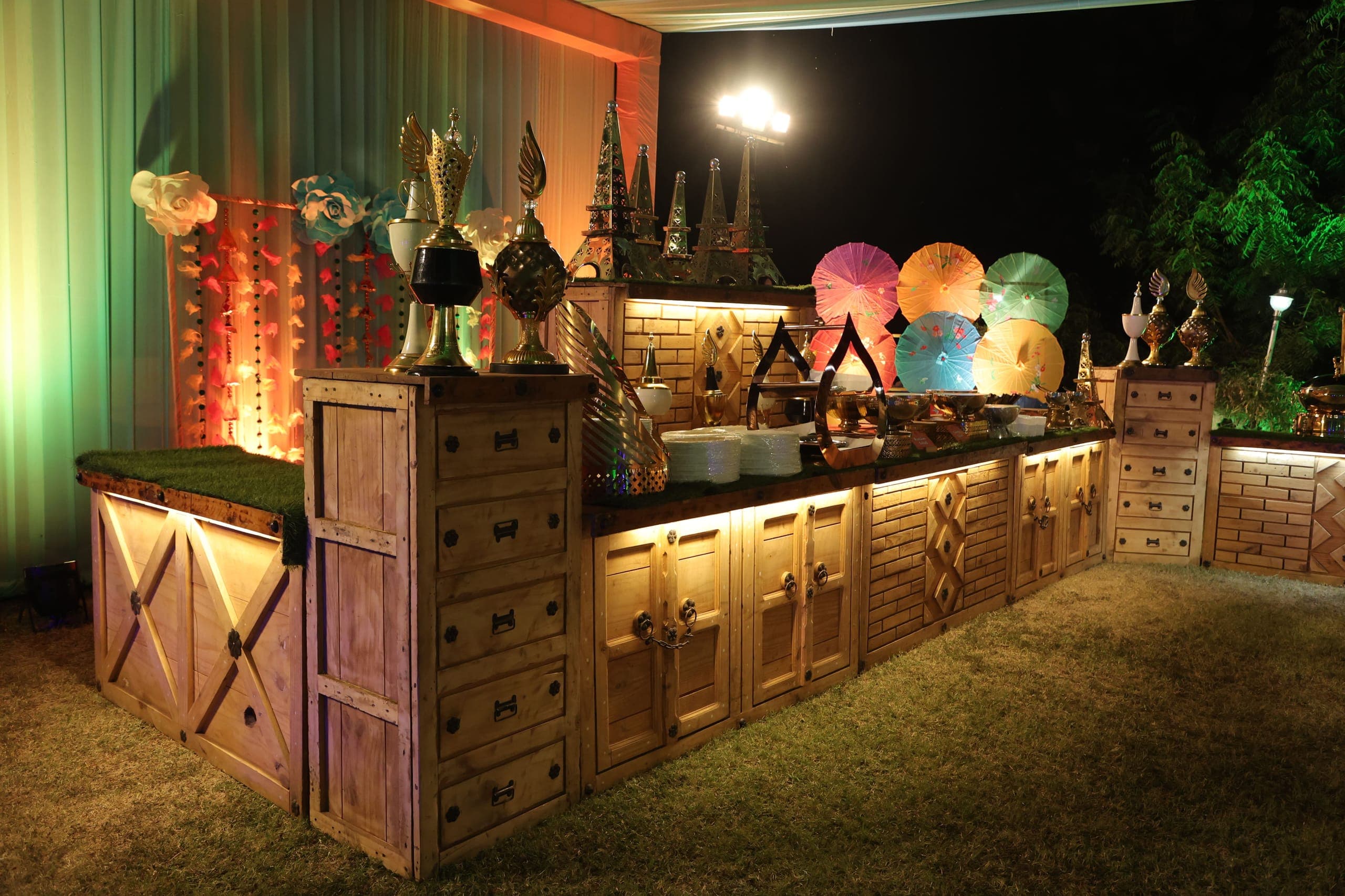 A long wooden buffet counter lit from beneath, set up on the lawn at night with brass serving dishes and painted paper parasols along the top