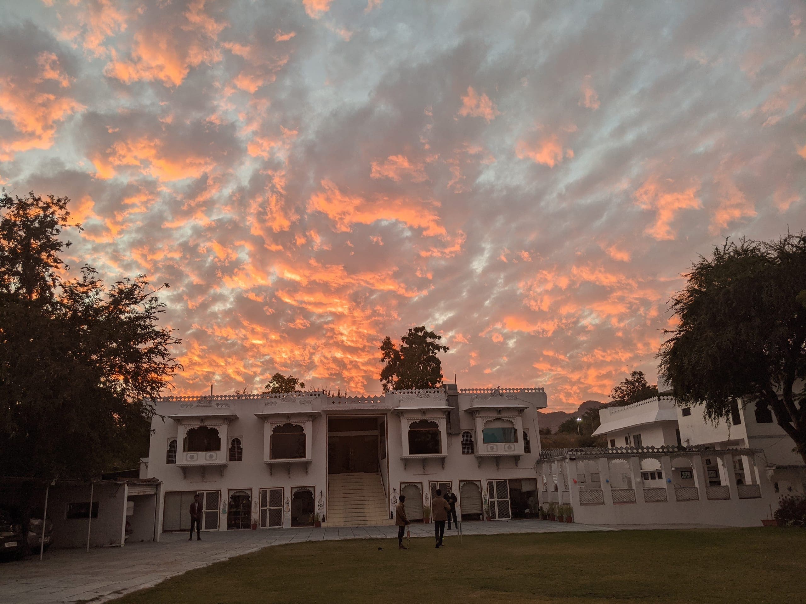 A white haveli with carved balconies seen from its lawn at dusk, under a sky of orange and grey cloud