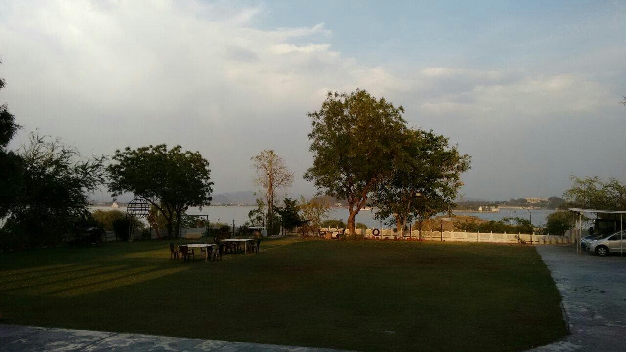 The lawn at Devendra Garh running down to Fateh Sagar in the late afternoon, with garden tables under a large tree and the far hills across the water